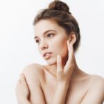 close-up-attractive-tender-young-european-woman-with-dark-hair-bun-hairstyle-being-naked-looking-aside-with-calm-expression-touching-face-with-hands-posing-magazine-photo-shoot